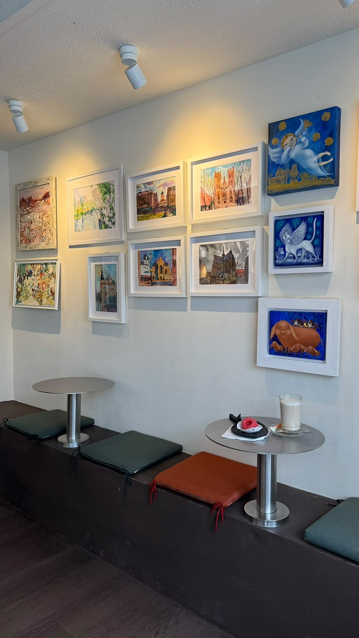 Gallery wall of framed paintings above a bench with cushions, small round tables, a latte and a raspberry dessert